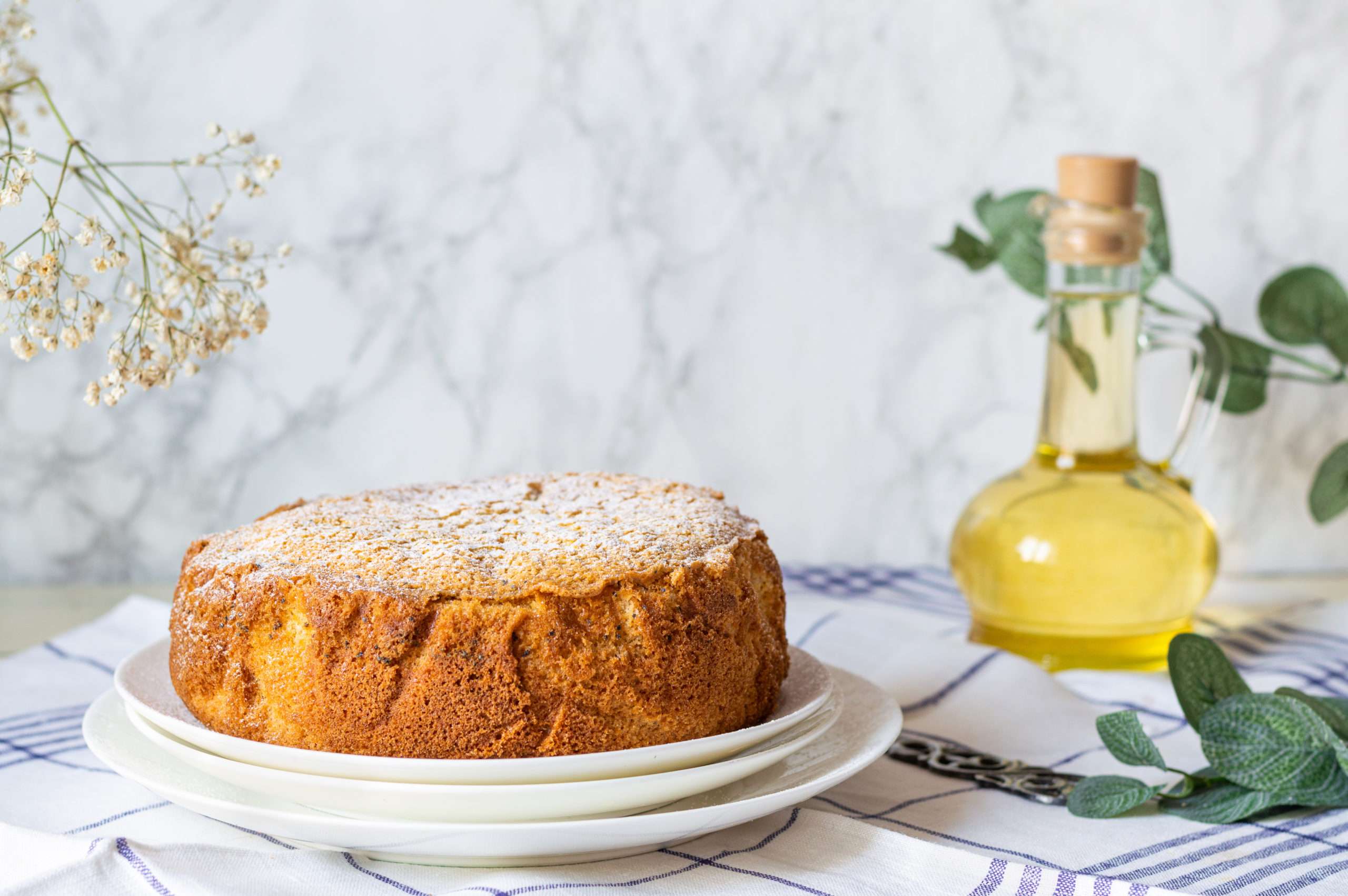 A classic olive oil cake sits elegantly atop a pristine white tablecloth adorned with blue stripes. Accompanying it, a bottle of premium olive oil stands nearby, while decorative green plants add a touch of freshness and charm to the scene.