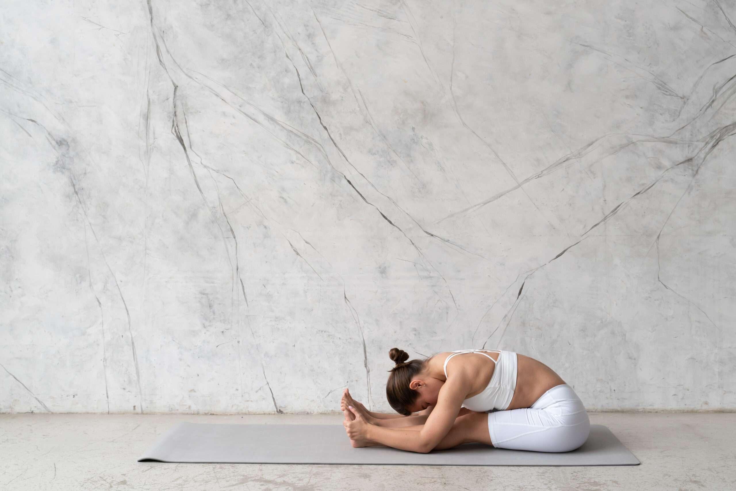 Young flexible woman practicing yoga in forward bend, paschimottanasana in trendy minimalist studio with a marble textured wall