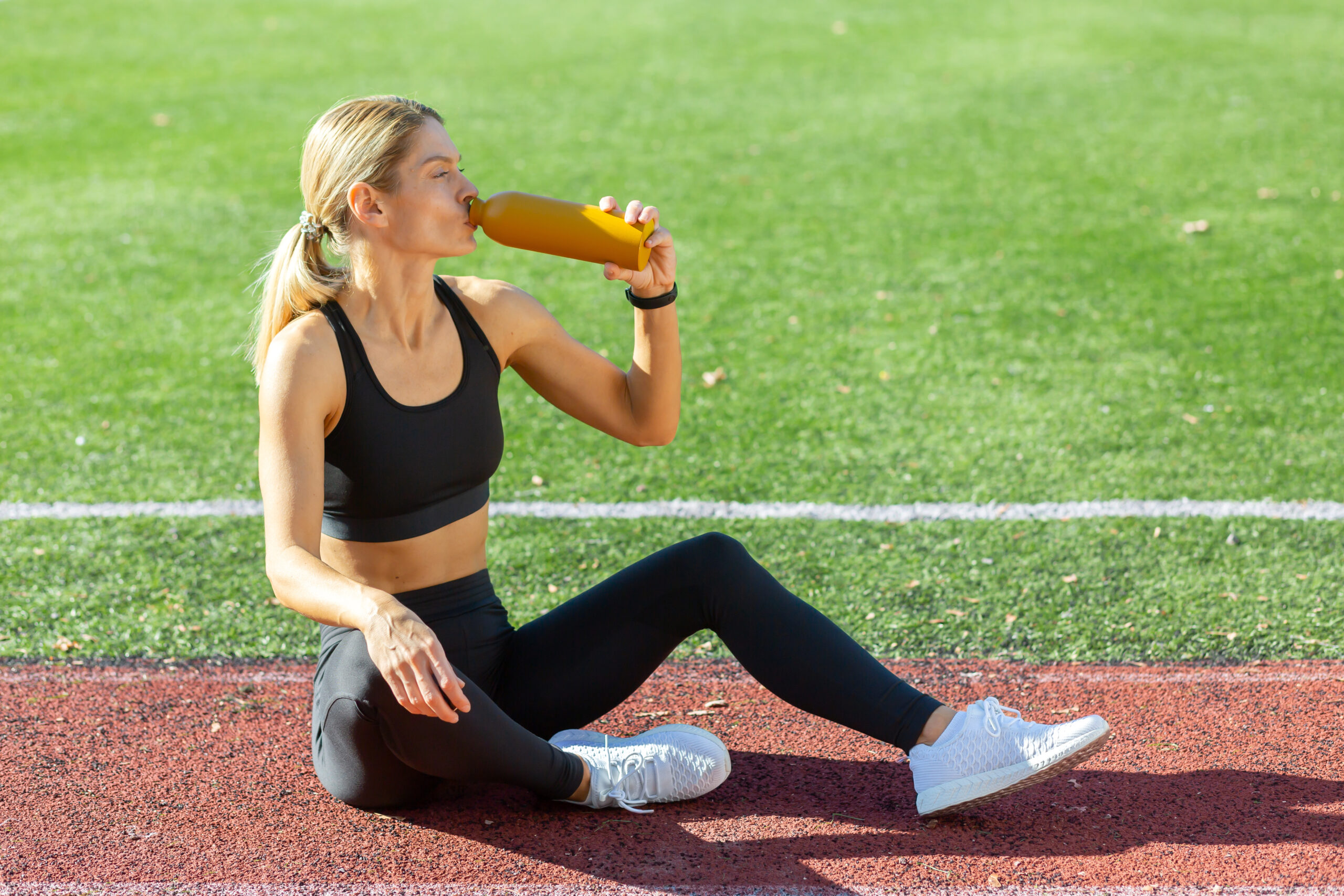 Blonde girl in black sports bra and black leggings sitting on a red track with green grass in the background drinking out of a orange reusable water bottle